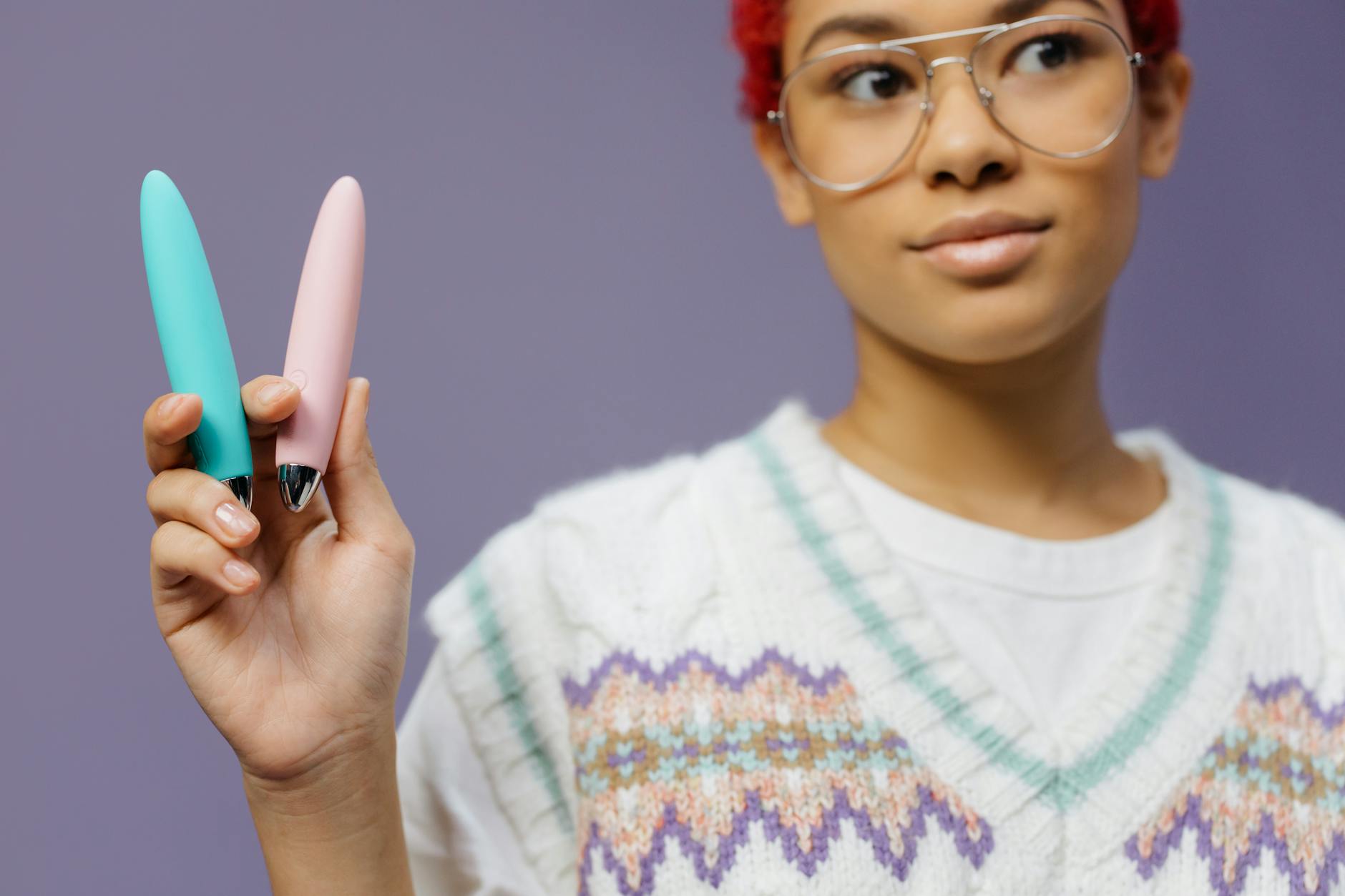 A woman holding two colorful vibrators against a purple background, expressing curiosity about exploring pleasure