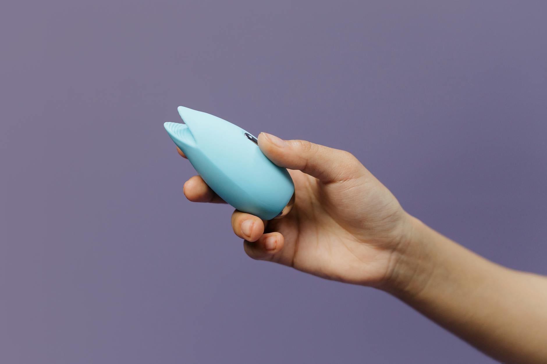 A blue silicone clitoral vibrator held in hand against a purple background